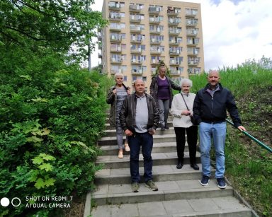 Na schodach kierownik Arkadiusz Wanat i seniorzy. Z boku zielone krzewy, z tyłu wieżowiec.