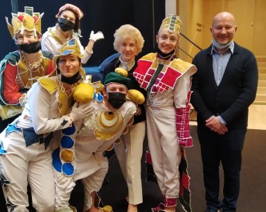 Sala teatru. Kierownik Arkadiusz Wanat, seniorka i aktorzy w kolorowych kostiumach stoją koło siebie uśmiechnięci.