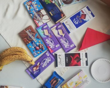 Na stole tabliczki czekolady w kolorowych opakowaniach. Obok czerwone serwetki, banany, papierowe talerzyki.