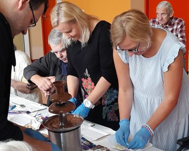 Przy stole pracownicy i seniorzy. Na blacie tabliczki czekolady w kolorowych opakowaniach, fontanna z płynną czekoladą.