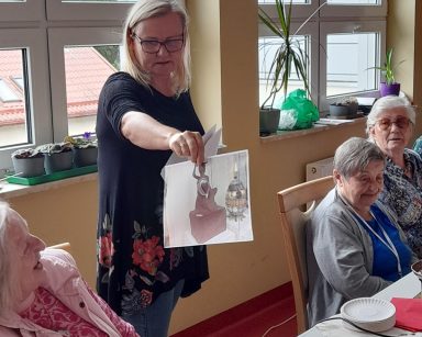 Przy stole seniorzy. Neurologopedka Ania Szmaja-Wysocka pokazuje zdjęcie syrenki z czekolady. Na blacie różne czekolady.