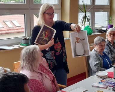 Przy stole seniorzy. Neurologopedka Ania Szmaja-Wysocka prowadzi zajęcia. Pokazuje zdjęcia. Na blacie różne czekolady.