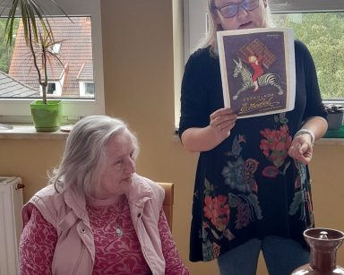 Neurologopedka Ania Szmaja-Wysocka pokazuje seniorom plakat z chłopcem na zebrze i tabliczkami czekolady na placach.