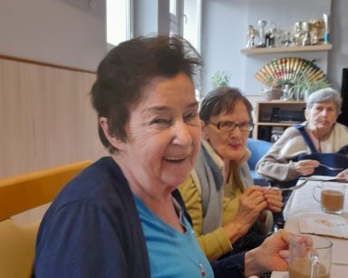 Trzy seniorki przy stole. Śmieją się. Przed nimi na blacie kubki z kawą.