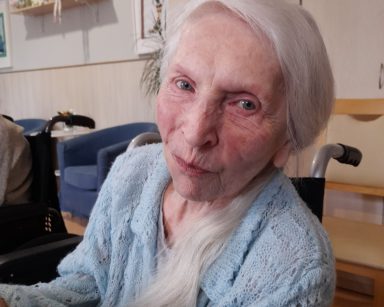 Jasny pokój. Seniorka siedzi uśmiechnięta.