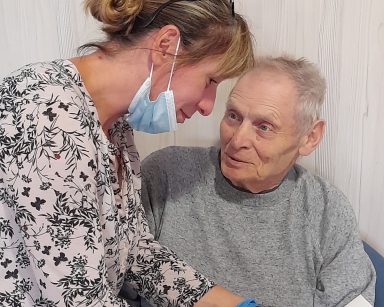 Kierowniczka Ewa Siłakiewicz-Witt nachyla się nad seniorem. W dłoniach trzyma tabliczkę czekolady. Senior uśmiecha się.