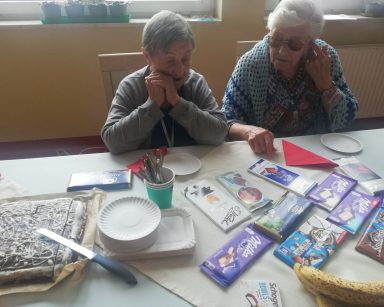 Dwie seniorki przy stole. Na blacie różne czekolady, czekoladowe ciasto, banany, papierowe talerzyki.