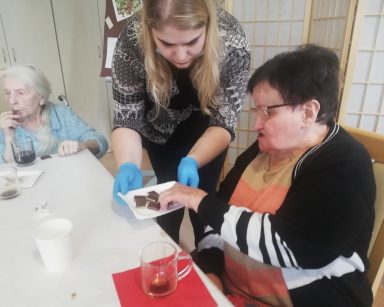 Przy stole seniorki. Pracownik częstuje je kostkami czekolady. Na blacie kubki z kawą.