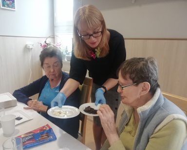 Przy stole dwie seniorki. Dyrektorka Agnieszka Cysewska nachyla się nad nimi i częstuje je czekoladą.