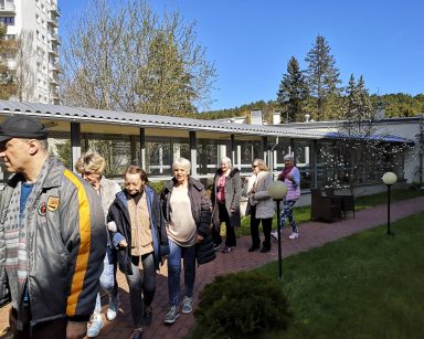 Słoneczny dzień. Kwitną kwiaty i drzewa. Seniorzy spacerują po ogrodzie.