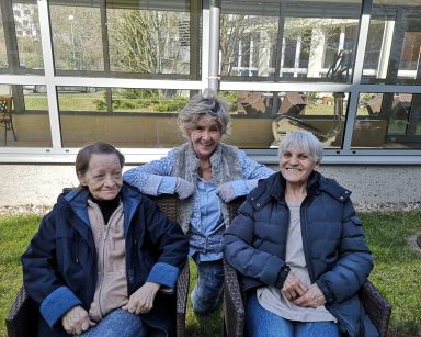 Ogród. Słoneczny dzień. Na fotelach siedzą dwie seniorki. Trzecia seniorka nachyla się do nich. Śmieją się.