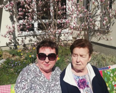 Słoneczny dzień. W ogrodzie na ławeczce siedzi seniorka i kierowniczka Mariola Ludwicka. Z tyłu kwitnąca magnolia.