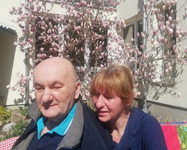 Słoneczny dzień. Ogród. Na ławeczce kierowniczka Ewa Siłakiewicz-Witt obejmuje seniora. Z tyłu kwitnąca magnolia.