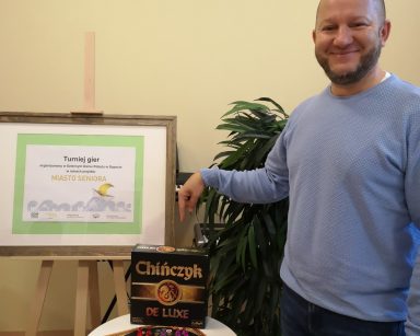 Sala. Kierownik Arkadiusz Wanat stoi przy stoliku. Uśmiecha się. Wskazuje na grę 
