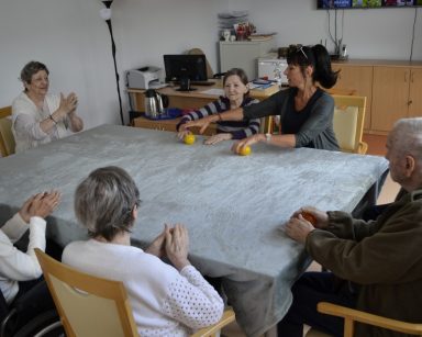 Sala. Przy stole siedzi terapeutka Ania Rzepczyńska i seniorzy. Ćwiczą ręce.