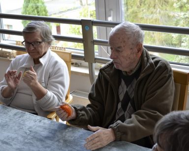 Sala. Przy stole siedzą seniorzy. Ćwiczą ręce za pomocą piłeczek.