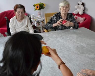 Sala. Przy stole siedzi terapeutka Ania Rzepczyńska i seniorzy. Ćwiczą ręce.