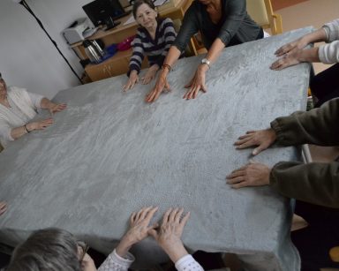 Sala. Przy stole siedzi terapeutka Ania Rzepczyńska i seniorzy. Ćwiczą ręce.