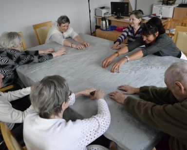 Sala. Przy stole siedzi terapeutka Ania Rzepczyńska i seniorzy. Ćwiczą ręce.