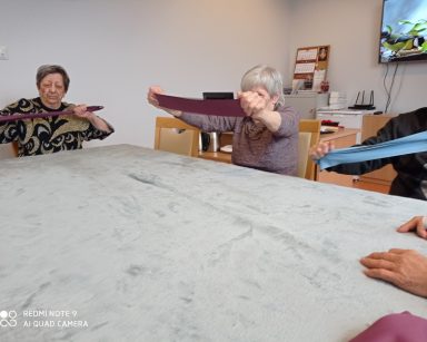 Sala. Przy stole siedzą seniorzy. Ćwiczą ręce z taśmami.