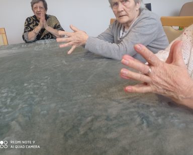 Sala. Przy stole siedzą seniorzy. Ćwiczą ręce.