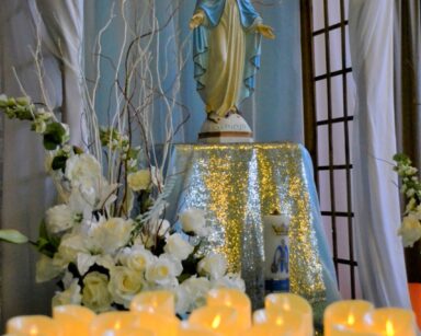Sala. Na podwyższeniu figurka Matki Boskiej. Przed nią ustawione zapalone świeczki.
