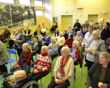 Sala. Na sali duża grupa seniorów i pracowników. Część z nich stoi, inni siedzą. Niektórzy trzymają zapalone świeczki.