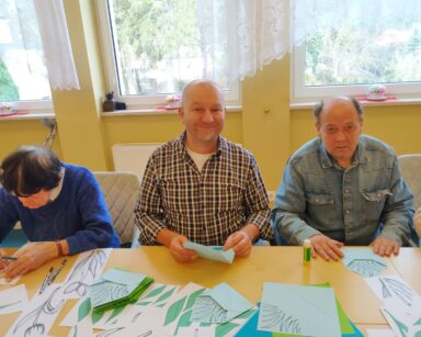Zbliżenie. Stół. Przy stole trzy osoby kolorują i wycinają tulipany. Na stole kolorowanki, nożyczki, kredki.