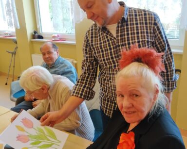 Sala. Mężczyzna trzyma w ręku kartkę z kolorowymi kwiatami. Obok seniorka pozuje do zdjęcia, w ręku trzyma kredki.