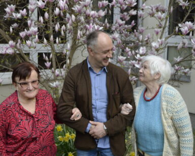 Ogród. Drzewo magnolii. Przy drzewie dwie seniorki trzymają pod rękę mężczyznę.