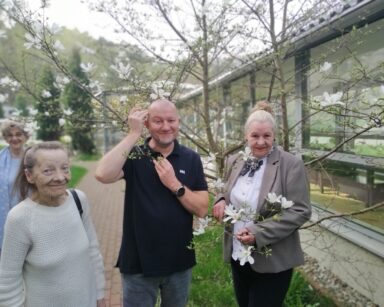 Ogród. 3 seniorki i mężczyzna pozują do zdjęcia. Mężczyzna stoi pod kwiatami magnolii, obok seniorka trzyma gałązkę drzewa.