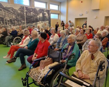 Sala. Na sali siedzi duża grupa seniorów, przy drzwiach stoją opiekunowie.