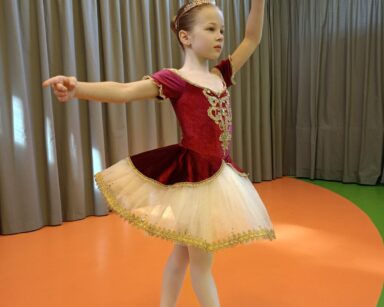 Zbliżenie. Na scenie mała baletnica w czerwono-białej sukience. Na głowie korona, na nogach jasne rajstopy i baletki.
