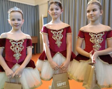 Zbliżenie. Trzy baletnice w czerwono białych sukienkach pozują do zdjęcia. W dłoniach trzymają torebki z napisem Sopot.