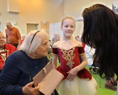 Zbliżenie. Mała baletnica stoi przy seniorce na wózku. Obok kobieta w czarnych włosach pochyla się do seniorki.
