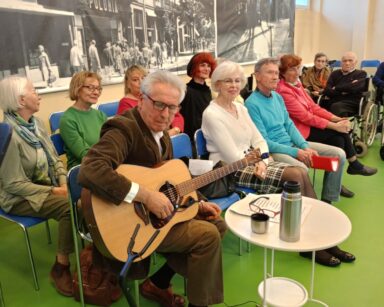 Zbliżenie. Pod ścianą rząd krzesełek, siedzi grupa ludzi. Mężczyzna trzyma gitarę, przed nim stolik, notatki i okulary.