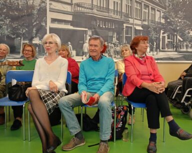 Sala. Rząd krzesełek pod ścianą, grupa ludzi siedzi. Kobieta w białej bluzce i spódnicy w kratę pozuje do zdjęcia.