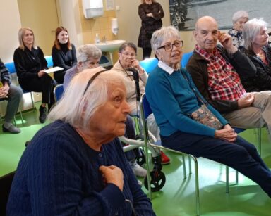 Sala. Grupa seniorów siedzi na krzesłach. Zbliżenie na seniorkę w granatowym swetrze, która palcem wskazuje na siebie.