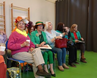 Sala. Rząd krzeseł pod ścianą, na nich siedzą barwnie ubrane seniorki. Jedna na głowie ma kolorowy, duży wianek.