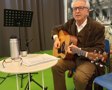 Zbliżenie. Mężczyzna w brązowym garniturze trzyma gitarę, siedzi na krześle, Przed nim na stoliku tekst, termos i okulary.