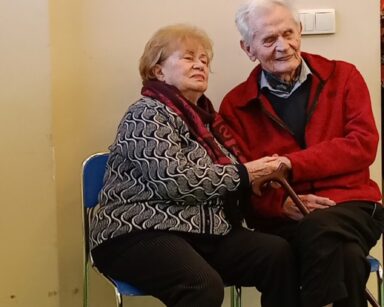 Zbliżenie. Para seniorów siedzi na krzesłach. Para trzyma się za ręce i słucha występu.