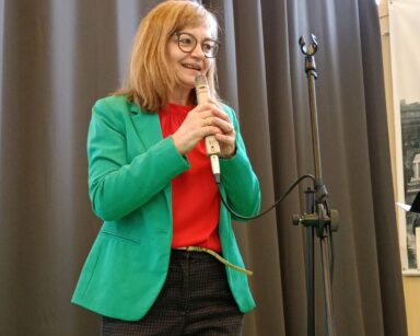 Zbliżenie. Kobieta w zielonej marynarce, czerwonej bluzce i spodniach w kratę trzyma w ręku mikrofon.