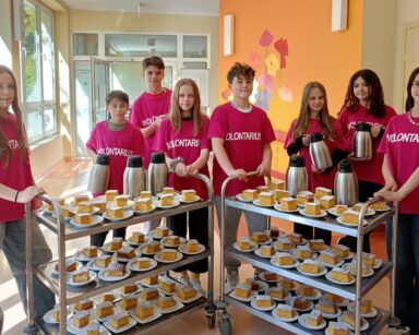 Sala. Grupa młodych wolontariuszy w różowych koszulkach z napisem pozuje do zdjęcia przy wózkach z ciastem i kawą.