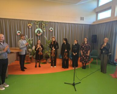 Sala. Na scenie grupa ludzi, w tym kilkoro muzyków trzymających w rękach skrzypce.