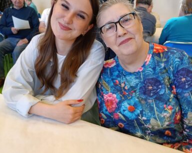 Zbliżenie. Młoda kobieta w ciemnych włosach i jasnej bluzce, pozuje do zdjęcia z seniorką w blond włosach i kolorowej bluzce.