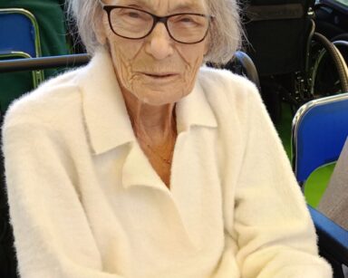 Zbliżenie. Uśmiechnięta seniorka w jasnym swetrze, siwych półdługich włosach, pozuje do zdjęcia.