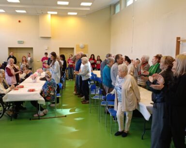 Sala. Przy stołach stoją seniorzy i młodzi ludzie, bija brawa i śpiewają. Na stołach kwiaty i pudełka czekoladek.