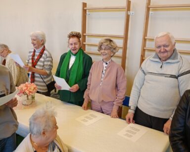 Zbliżenie. Pięcioro seniorów stoi przy stole, na stole karta z piosenką. Seniorzy śpiewają i uśmiechają się.