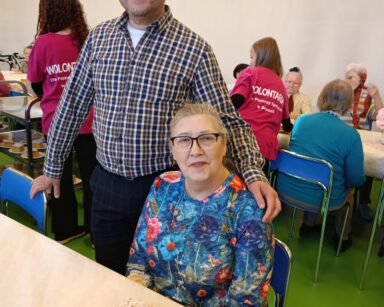 Zbliżenie. Mężczyzna trzyma dłoń na ramieniu uśmiechniętej seniorki, która siedzi przy stole. Para pozuje do zdjęcia.