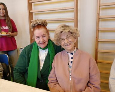 Zbliżenie. Dwie uśmiechnięte seniorki, ubrane na kolorowo, pozują do zdjęcia przy stole. W tle wolontariuszka niesie ciasto.